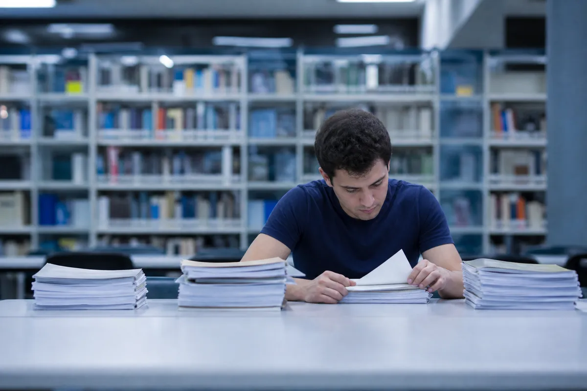 Estudante numa mesa de biblioteca com quatro pilhas de material de estudo separadas por área do conhecimento, folheando uma delas