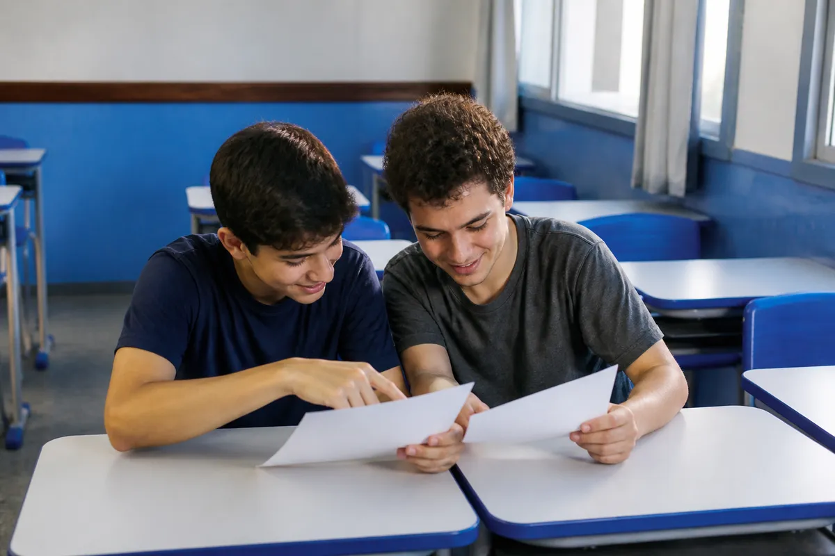 Dois estudantes numa sala de aula comparando duas folhas de redação sobre a carteira, um deles apontando um trecho